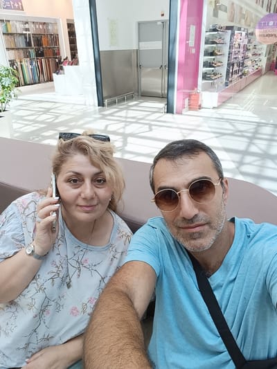 A man and a woman are seated on a brown bench in what appears to be a shopping mall in Novaya Adygeya, Russia. The woman, on the left, has blonde hair and is wearing a light blue floral print shirt. She is holding a smartphone to her ear and appears to be in conversation. She is also wearing a watch and rings on her fingers. The man, on the right, has short grey hair and is wearing a light blue t-shirt. He has on round, gold-rimmed sunglasses. Behind them, shelves stocked with various items are visible, including what looks like fabric samples and makeup products. The floor is tiled, and sunlight casts geometric shadows across the area. A bright pink wall and a stainless steel door are also part of the background.