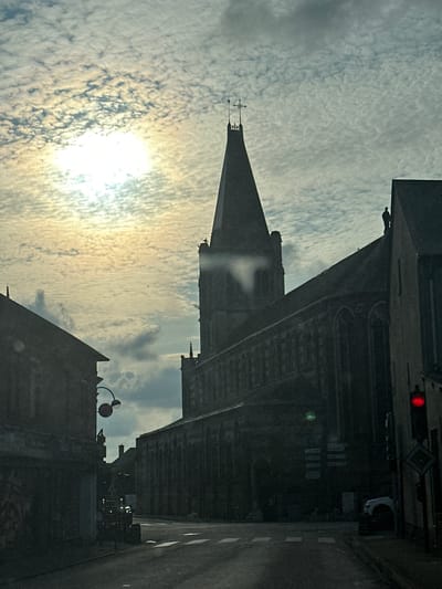 The image depicts a street scene in Saint-Maclou-de-Folleville, France, dominated by the silhouette of a large church with a tall spire. The sun is low in the sky, partially obscured by textured clouds, casting a diffused light that creates a somewhat muted and dramatic atmosphere. In the foreground, the dark facades of buildings line the street, one of which appears to be a shop with faded lettering. The road in front of the church has crosswalk markings and appears wet or reflective. A red traffic light is visible on the right side of the frame. There is a white car parked on the right side of the road, and a vehicle that could be a truck or van is visible further down the street in the center. A person is visible on the roof of a building to the right of the church spire, and another figure stands near the top of the church's spire. Signposts are affixed to the wall of the church.