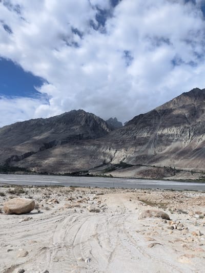 This image captures a vast, arid landscape under a partly cloudy sky. In the foreground, a sandy, uneven track is visible, marked by tire treads, with scattered rocks and sparse vegetation. A large, smooth boulder rests on the left side of the track. Beyond the track, a wide, dry riverbed or plain stretches out, appearing dusty and desolate. In the mid-ground, a line of trees and some buildings can be seen along the edge of the riverbed, indicating a settlement or village. The background is dominated by imposing, rugged mountains with steep, eroded slopes. The mountains display a range of textures and colors, from dark grays and browns to lighter sandy hues, with shadows accentuating their contours. The sky above is a mix of bright blue patches and large, dramatic clouds, some of which are dark and heavy, suggesting possible rain or a change in weather. The overall impression is one of a remote, mountainous region, characteristic of areas like Sasoma, India, known for its stark natural beauty and challenging terrain.