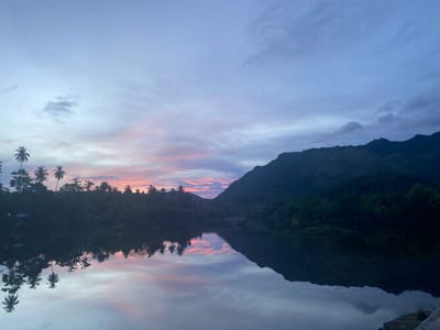 The image captures a serene twilight scene over a body of water in Aneuk Paya, Indonesia. The sky is a gradient of pale blue and soft pink hues, reflecting in the calm water below. Silhouetted palm trees and lush green vegetation line the left bank of the water, their reflections mirrored perfectly on the surface. To the right, a dark, imposing mountain range dominates the horizon, also partially reflected in the water. The overall atmosphere is tranquil and natural.