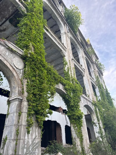 The image shows a low-angle view of an abandoned, overgrown building in Tsanbaikwara, Abkhazia. The building features a repeating pattern of arched openings and rectangular architectural elements. Heavy vines and vegetation cover large portions of the facade. The structure is weathered and shows signs of decay. Some sections of the building are missing portions of walls. The sky is visible above the building and is a mixture of light blue and white clouds.