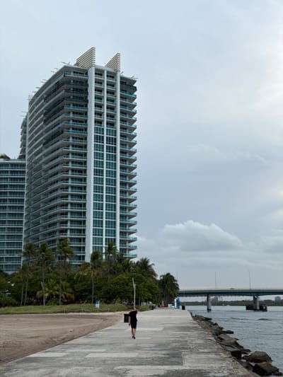 A full shot captures a person walking on a concrete pier, facing away from the camera, with a large, modern building in the background. The building, featuring numerous balconies and glass windows, is tall and appears to be under construction in some sections. Palm trees are scattered around the base of the building, and a beach is visible to the left. The pier extends towards a body of water on the right, where a bridge spans across it in the distance. The sky is overcast, suggesting a cloudy day. The scene is set in Bal Harbour Village, United States of America.