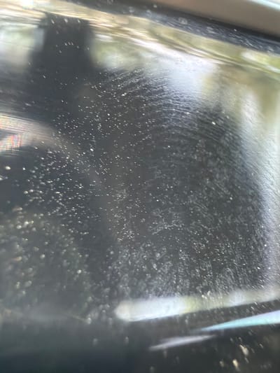 This is a close-up, abstract image of a dark, textured surface, possibly the interior of a car or a piece of equipment. The surface is covered in numerous small, white specks that appear to be dust or debris. There are also visible water droplets or condensation, creating a streaky, textured pattern across the surface. Light reflects off the surface in various ways, creating bright white highlights and subtle rainbow-like refractions, suggesting an artificial light source or sunlight filtering through. The overall impression is one of a slightly grimy or neglected surface. There is no discernible activity, people, or specific setting beyond the textural details of the surface itself. The image is abstract and lacks clear context for time of day, weather, or location beyond what can be inferred from the reflective qualities of the material. There is no visible text.
