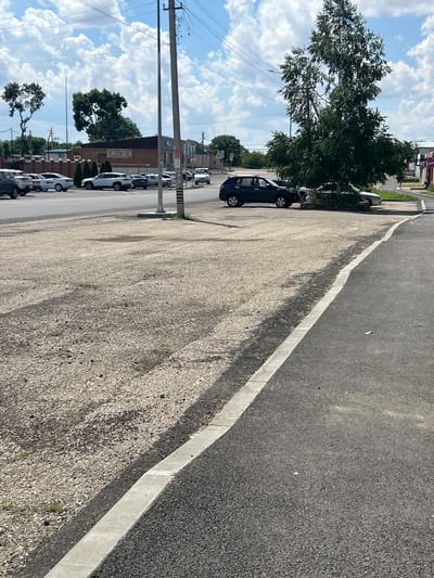 This media file depicts a street scene in Dondukovskaya, Russia. The scene is dominated by an asphalt road with a white line marking its edge, bordering a gravel shoulder. Several cars are parked along the gravel area, including a dark-colored hatchback in the foreground. Trees, some with dense foliage, line the opposite side of the street, interspersed with buildings. Power lines and utility poles are also visible, suggesting an inhabited area. The sky is partly cloudy, with large, white cumulus clouds against a bright blue background, indicating it is daytime with fair weather. The overall impression is one of a typical, somewhat mundane day in a Russian town, with no specific activity or event clearly taking place beyond the presence of parked cars. There are no discernible people in the image, and no visible text that can be read. The lighting suggests it is likely midday or early afternoon.