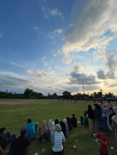 In the city of Aratontonmoncrang, Indonesia, a soccer match unfolds on a grassy field under a dynamic sky filled with scattered clouds. The setting sun casts a warm glow, illuminating the scene with a soft, golden light. Spectators, a mix of men, women, and children, are gathered on the field's edge, some sitting on the grass, others standing. Many of the women are wearing hijabs. In the background, a row of trees and buildings forms a natural backdrop, with a telecommunications tower standing tall among them. Players in red jerseys are visible on the field, engaged in the game. The overall atmosphere is one of community engagement and leisure activity.