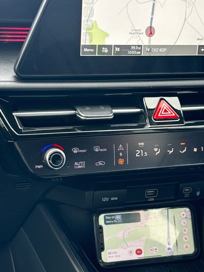 The image captures the interior of a car, focusing on the dashboard and center console. On the left, the car's built-in infotainment screen displays a navigation map, showing the current location relative to a destination. Visible text on this screen includes "Creech Heathfield," "39.3 mi" remaining distance, a destination postcode "EX2 8QP," and a time of "10:03 AM." The map also shows a "Menu" option and a scale of "Auto 0.25 mi."

In the center, a vertical panel features climate controls and air vents. A red triangular hazard light button is prominent. The digital climate display shows "SYNC" and a temperature of "21.5" degrees Celsius. Below this are buttons for air circulation, fan speed, front and rear window defrost ("FRONT," "REAR"), and an "AUTO CLIMATE" setting, with a "PWR" knob.

To the right, a smartphone, likely an iPhone, is resting in a storage or charging area, also displaying a navigation app. This app shows "Stay on M5," "36mi," and "42 min" estimated travel time. Adjacent to the phone are "USB" ports and a "12V 180W" power outlet.

The scene suggests a vehicle in transit, likely during the morning, given the 10:03 AM time. Both the car's navigation and the smartphone's navigation are active, indicating an ongoing journey in the United Kingdom, specifically referencing the M5 motorway and a UK postcode. The climate control is set to a moderate temperature, suggesting a comfortable interior environment.