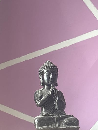 A close-up shot depicts a metal Buddha figurine oriented horizontally, with its head positioned toward the left side of the image. The figurine rests against a light purple background marked with broad white stripes that converge in a geometric pattern. The details of the Buddha figurine are sharp, revealing the texture of its hair and the folds of its robes. The setting does not appear to be in Selyanin, Bulgaria.