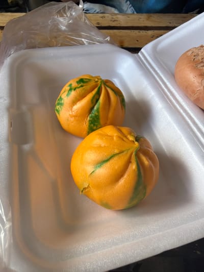 This image captures a close-up view of food items packaged in a white styrofoam container, likely for takeout or sale in Măng Đen, Vietnam.

The primary subjects are two brightly colored buns, appearing to be steamed buns (like *bao* or *bánh bao*). They are predominantly orange with striking, irregular green streaks and patches, creating a marbled or patterned effect. Both buns are round and plump, with visible folds in their dough, particularly at the top where they seem to have been sealed.

Partially visible in the upper left corner of the container is a third bun or piece of bread, which is a uniform light brown color and has a rough, textured surface, different in appearance from the orange and green buns.

The buns are neatly placed within the white styrofoam container, which shows some wear and minor indentations. To the left of the container, a clear, crinkled plastic bag is visible, partially obscured by the container's edge. Below and to the far left, a wooden surface, possibly a table or crate, with a rustic grain and dark specks, serves as the background.

The scene depicts food ready for consumption or purchase. The style of buns and packaging are consistent with street food or market offerings common in Vietnam. There are no people visible, no discernible activity beyond the presentation of the food, and no visible text. The lighting is soft and even, suggesting either an indoor setting or a shaded outdoor area.