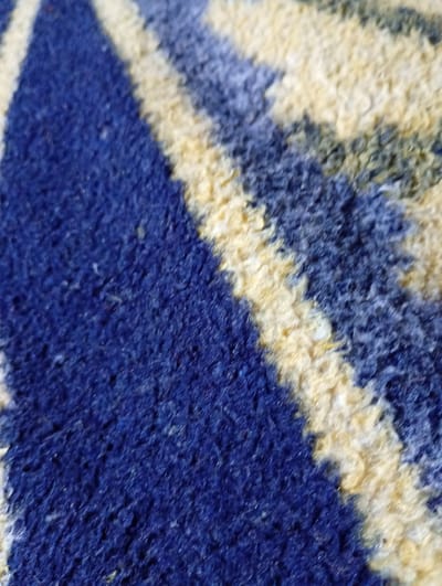 A close-up, high-angle shot captures a section of a blue and yellow striped carpet. The carpet fibers are thick and appear plush, with the blue sections being a deep, rich shade and the yellow sections a lighter, more muted tone. The stripes run diagonally across the frame, creating a sense of movement. The focus is soft, giving the image an abstract feel. There is no discernible information about the location in Lanchkhuti, Georgia, nor are there any people, actions, or interactions depicted in the image.