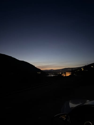 The image depicts a panoramic view of a valley at twilight or early night, situated in the city of Lamego, Portugal. The sky transitions from dark navy blue overhead to a faint orange and yellow glow at the distant horizon, indicative of a sunset or sunrise. Silhouetted hillsides flank the scene to the left and right, their dark forms contrasting with the sky. Scattered artificial lights are visible on these hills, indicating distant structures or settlements.

In the mid-ground, a dark, reflective body of water, likely a river, winds through the valley. A prominent, square-shaped building, possibly a tower or part of a larger structure, stands illuminated in the mid-right, its walls glowing light yellow-orange. Further in the distance, a bridge spans across the valley, marked by a line of artificial lights. Beyond this, numerous small, diffused lights delineate a distant town or settlement stretching across the valley and up the opposing hills.

The foreground shows the interior of a vehicle. The dark, curved top of a steering wheel is visible on the bottom right, with a faint circular emblem at its center. Part of the vehicle's dashboard or hood is also visible, reflecting minimal light. A dark, flat surface, appearing to be a road or a section of railroad tracks, runs across the lower mid-ground, leading towards the illuminated distant landscape. No people or explicit actions are discernible.