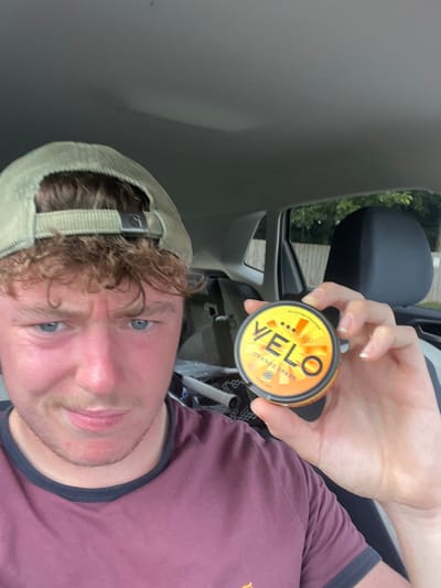 A forensic description of the scene reveals a single young adult male seated inside a vehicle, likely in Cherry Burton, United Kingdom. The individual has reddish-brown, curly hair, blue eyes, and wears a light olive-green corduroy baseball cap backward and a maroon t-shirt with a dark blue collar. His facial expression is one of apparent displeasure, characterized by furrowed brows and slightly pursed lips. He holds a circular orange and yellow container, identified as "Velo" brand nicotine pouches, labeled "Orange Spark" with a four-dot strength indicator and "Made in UK" text, in his right hand, positioned towards the right side of the frame. The vehicle's interior features a light-colored ceiling and dark grey or black seating. Through the vehicle's rear windows, green foliage, consistent with trees or bushes, and a light-colored fence are visible. Several white cylindrical objects and other miscellaneous items are stored in the vehicle's rear cargo area.