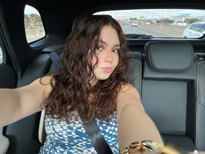 A woman with curly brown hair and a blue and white patterned dress is sitting in a car. She is wearing a seatbelt. Her head is tilted back, and she is looking at the camera. The view out the window shows a highway with cars. The car's interior is black. A gold watch is visible on her wrist.