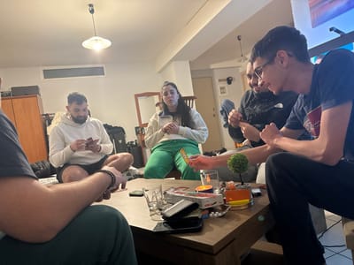 The image depicts five individuals in an indoor residential setting in Plataniás, Greece, engaged in a card game around a rectangular wooden coffee table.
The environment features light-colored walls, an overhead ceiling light fixture with a white shade, a high horizontal window, a large wooden wardrobe on the left, and a mirror over a dresser on the right side of the background. A television screen is partially visible on the far right.
The individuals are:
1. A male subject with his back to the camera in the immediate foreground, wearing a dark short-sleeved shirt and dark pants, with a dark beaded bracelet on his left wrist.
2. A male subject seated on the floor to the left, cross-legged, wearing a light-colored hooded sweatshirt and dark shorts, holding and examining a hand of cards.
3. A female subject seated in the background-center, wearing a light-colored zippered top and green pants, with her hands resting clasped.
4. A male subject to the right-background, partially obscured, wearing a dark jacket with a white logo, holding a small card or object.
5. A male subject with glasses in the right-foreground, wearing a dark t-shirt with a white graphic, extending his left hand to display a green and orange card, while looking towards the center of the table.
The coffee table contains multiple objects: several clear drinking glasses, a small artificial green topiary plant, a lit candle in a metallic holder, an orange container, a dark-cased smartphone, small white circular objects on a wrapper, and various card game components.