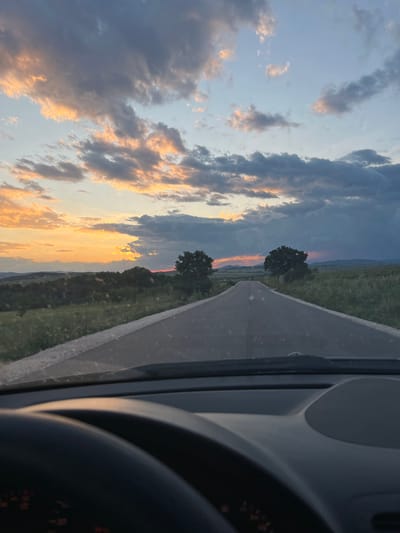 The image shows a view from inside a car on a road, likely in transit. A gray road stretches out, bordered by green fields. Two prominent trees stand near the road. The sky is filled with clouds illuminated by the setting sun, displaying a range of colors from orange to gray. Parts of the car interior are visible, including the dashboard and steering wheel. The image is taken from the city of Smilets, Bulgaria.