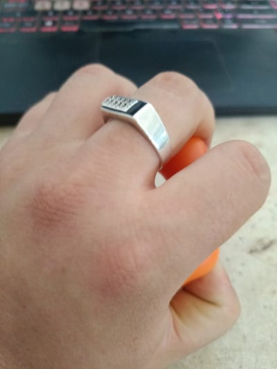 A close-up, horizontal, eye-level shot captures a person's hand, adorned with a silver ring, gripping an orange object. The ring features a textured, black rectangular inlay. The background is blurred, showing a dark surface with hints of red and pink, likely a keyboard. The image is taken indoors, possibly in a domestic or office setting, with no discernible connection to Berdiansk, Ukraine. The focus is on the hand and the ring, with the surrounding environment out of focus.