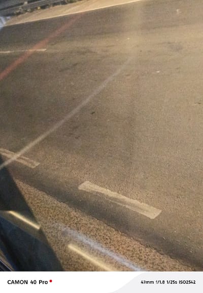 The image presents a low-light view of an asphalt road or paved surface, likely captured at night or during dusk from a slightly elevated perspective, possibly from inside a vehicle. The road surface is dark grayish-brown, displaying a granular, textured appearance.

Prominent features on the road include several white markings and distinct light reflections. A long, continuous white line runs diagonally from the bottom right towards the top left of the frame, appearing brightly illuminated. Additionally, several short, rectangular dashed white lines are visible, scattered across the mid-left and bottom-right sections, some appearing faded or worn. A faint reddish-orange streak of light or reflection runs diagonally across the upper-middle portion of the asphalt, roughly parallel to the main white line.

The scene is characterized by striking linear light reflections. A bright, diffuse white light streak extends from the bottom left corner, traversing both the asphalt and a darker, angular object that occupies the lower left quarter of the frame. This object appears to be a dark, textured, and somewhat reflective surface, possibly a curb, a barrier, or an interior component of the vehicle from which the photo was taken. Other subtle reflections are also present on this dark surface.

The overall lighting is dim, contributing to a noticeable amount of digital noise in the image, especially visible in the uniform areas of the asphalt. This is consistent with the indicated exposure settings.

Visible text overlays at the bottom of the image provide technical details:
*   "CAMON 40 Pro" - identifying the camera model used.
*   "47mm f/1.8 1/25s ISO2542" - indicating the photographic exposure parameters (focal length, aperture, shutter speed, and ISO sensitivity). The high ISO value of 2542 confirms the low-light shooting conditions.