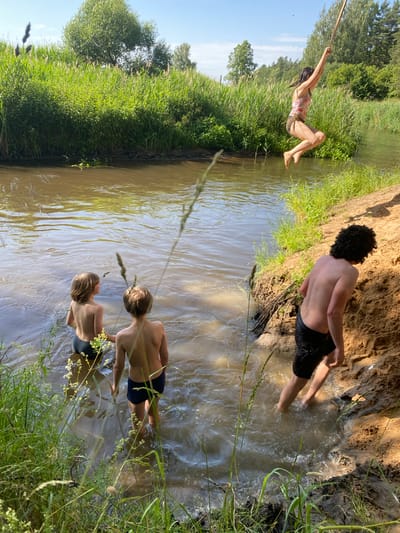 The image depicts a sunny day scene at a riverbank in Klyontski Sel'ski Savet, Belarus. Three young individuals are enjoying the water. In the foreground, two boys are wading in the shallow, muddy water. One boy, facing away from the camera, has his legs submerged and is looking towards the river. The other boy, also facing away, is slightly further out in the water, with his back visible. In the background, another person, possibly a girl, is swinging from a rope over the water, her body arched backward and her arms extended. She appears to be mid-swing, with her feet close to the water's surface. The river is a murky brown, reflecting the bright sunlight. Lush green reeds and grass line the banks, with trees visible in the distance under a clear blue sky. The overall atmosphere is one of carefree summer fun and outdoor recreation. There is no visible text in the image.