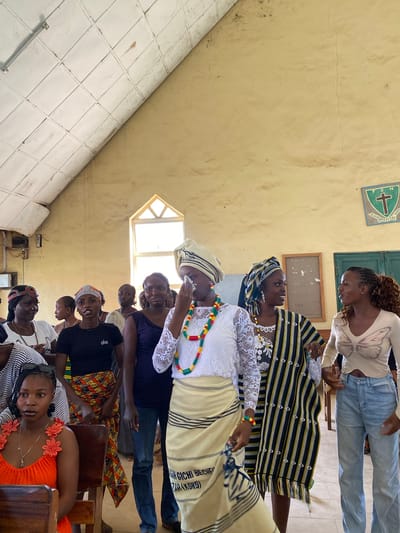 This image captures a gathering of people, likely women and girls, in an indoor setting. The scene appears to be a community space with cream-colored walls and a tiled floor. Sunlight streams in from windows, suggesting it is daytime. Some individuals are dressed in traditional attire, featuring colorful patterns and headscarves, while others wear more casual modern clothing. One person in the foreground is wearing a bright orange top adorned with floral appliques and a necklace. A few people are looking towards the camera, and some seem to be engaged in conversation. There's a sense of community and possibly celebration or a cultural event taking place. A framed object hangs on the wall, and a small sign with a green cross and some text is also visible. The overall atmosphere seems warm and engaging.