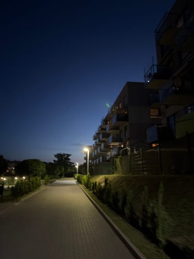 The image depicts a pathway lined with trees and bushes, illuminated by streetlights, leading towards a modern apartment building. The sky is a deep twilight blue, suggesting it is either early evening or late night. The building, with its balconies and glass railings, is silhouetted against the darkening sky. The pathway itself is paved with interlocking stones. The overall scene conveys a sense of quiet and order.