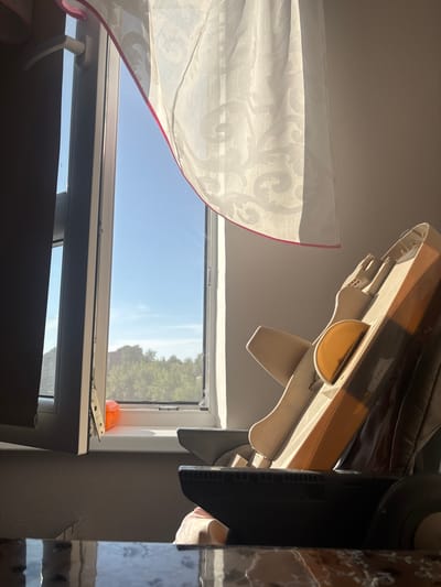 The image depicts an interior scene near a window. A light-colored curtain with a pink trim is partially drawn, allowing sunlight to stream in. The window itself offers a view of lush green trees and a clear blue sky. Inside, a child's high chair, primarily beige and orange, is positioned in the foreground. To the left of the window, a dark curtain is visible. The surface in the very foreground appears to be a reflective table with a dark, patterned top. The overall ambiance suggests a bright, sunny day. The specific location in Krāslava, Latvia, cannot be determined from the image.