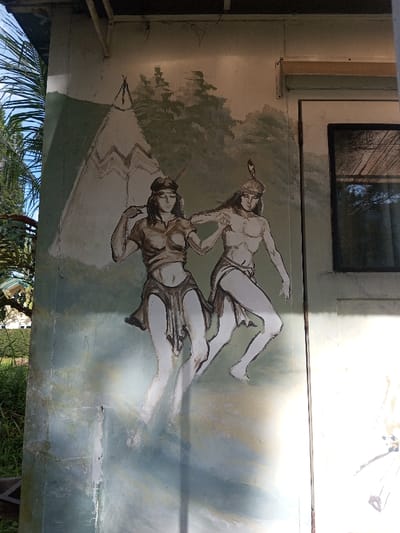 A faded mural graces the side of a building, depicting a Native American man and woman in traditional attire. They stand before a backdrop of a teepee and wooded hills. The colors are muted, with shades of white, brown, and green dominating the scene. The artwork is worn, with visible signs of aging and weathering. A nearby window suggests the mural is part of a building's exterior.