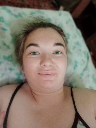 The image is a close-up, high-angle selfie of a young woman lying down. She has light skin, medium-toned hair that appears to be light brown or dirty blonde, and bright green eyes. Her eyebrows are defined and dark. She is wearing a black tank top. The background consists of a light-colored pillow with a green floral pattern, and a darker, blurred surface behind it, possibly bedding or furniture. The scene appears to be indoors, in a domestic setting. The photographer is the subject of the image. There is no information in the image to suggest a specific location in Dybshche, Ukraine.