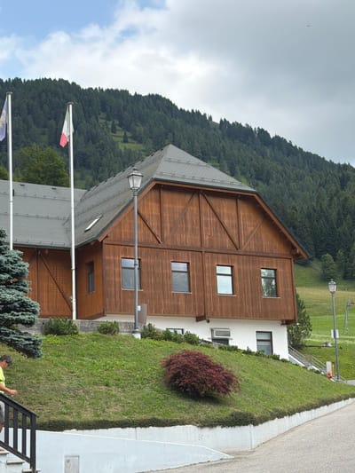 The image captures a view of a wooden building with a gray roof in Sauris / Zahre, Italy, set against a backdrop of a dense, green forest covering a hillside under a cloudy sky. Two flags, one of which appears to be the Italian flag, are visible on flagpoles in front of the building. A lamppost stands near the building, and a small, red bush is on a grassy slope. In the foreground, a person wearing a yellow shirt and shorts is walking down a set of stairs. The overall scene depicts an outdoor environment with a building and natural surroundings.