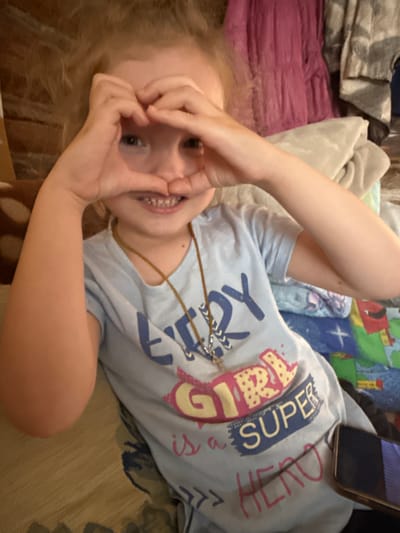 A young girl with blonde hair is sitting on a bed, making a heart shape with her hands over her face. She is wearing a light blue t-shirt with a graphic that reads "GIRL is a SUPER HERO". A necklace with a cross pendant is visible. The background includes pillows and blankets, suggesting a domestic setting. There is no identifiable information about the location in Severnyy, Russia.