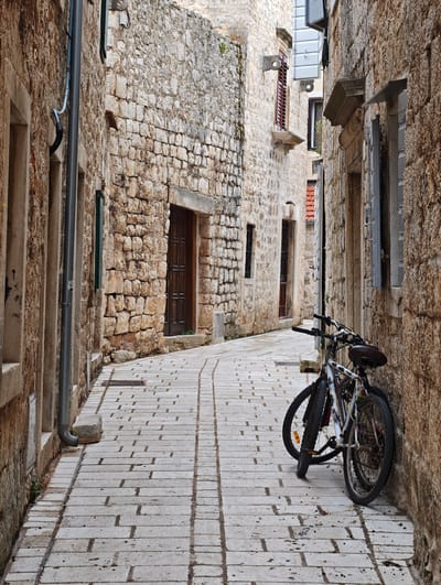 This is a street-level, eye-level, full-shot photograph taken in the city of Grad Stari Grad, Croatia. The scene depicts a narrow, cobblestone alleyway flanked by old stone buildings on both sides. The cobblestones are wet, suggesting recent rain, and exhibit a pattern of light and dark rectangular stones with visible grout lines.

On the right side of the alley, two bicycles are parked leaning against a rough-hewn stone wall. The bicycle in the foreground is white with colorful decals and has a dark saddle and handlebars. Its front wheel is turned sharply to the left, and its rear wheel is positioned to the right. The second bicycle is partially obscured behind the first, but its dark frame and wheels are visible.

The buildings are constructed from large, irregular stone blocks, giving them a rustic and historic appearance. The building on the left features a vertical gray downspout running down its facade, ending in a stone block support. There are windows and a dark wooden door visible on the buildings. The building on the right also displays a stone facade with a window above where the bicycles are parked. Shadows are cast on the cobblestone pavement, indicating indirect lighting. The overall atmosphere is one of quiet, ancient European urban life.