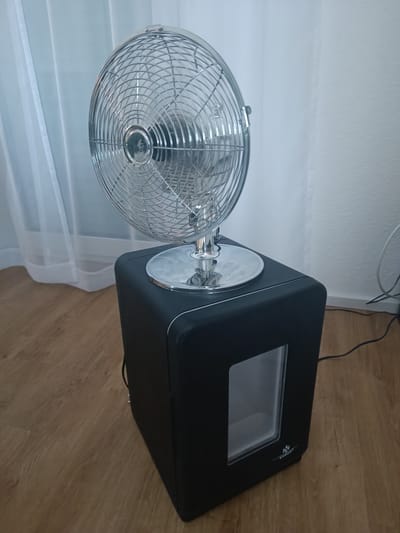 This is a media file featuring a retro-style chrome fan perched atop a small, black mini-fridge. The fan has a decorative grill and is mounted on a shiny chrome base. The mini-fridge is cubical with a clear window on its front, revealing a glimpse of its interior. A small logo, which appears to read "KESSER®", is visible on the lower right corner of the fridge. The scene is set indoors, on a light brown wooden floor. Behind the objects, sheer white curtains are visible, suggesting a window is present. The lighting appears to be natural, indicating daytime. The overall impression is one of a home or office space, possibly used for personal comfort or small-scale storage. There are no people present in the image. The image is shot from a slightly low angle, looking up at the fan and fridge. A black power cord is visible trailing from the back of the mini-fridge. The setting appears to be Germersheim, Germany.