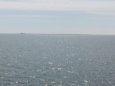 The image shows a wide expanse of water under a hazy sky. In the distance, a low, dark landmass stretches across the horizon. On this landmass, a few structures, including what appears to be a lighthouse or a tall building, are visible. The water's surface is rippled and reflects sunlight, creating a shimmering effect. The overall scene suggests an open, coastal environment. The location is identified as Kuivastu, Estonia, a port town on the island of Muhu, known for its ferry connections.