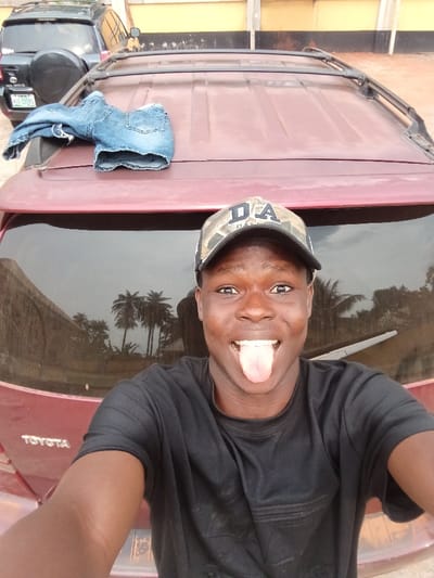 The image captures a young male taking a selfie outdoors in Itu, Nigeria. The subject, located in the foreground, wears a black t-shirt and a camouflage baseball cap displaying "DA" on its front, smiling widely with his tongue extended. He is positioned in front of a red/maroon Toyota SUV, identified by the partial "TOYOTA" logo on the lower left of the tailgate. A pair of folded light blue denim shorts rests on the SUV's roof, which also features a black roof rack. The SUV's tinted rear window reflects several palm trees and a cloudy sky. In the mid-ground, a dark-colored SUV with a rear-mounted spare tire is visible, bearing a Nigerian license plate "KGC 155 HH", with a passenger door ajar. The background features a light yellow/tan building wall with a darker base and horizontal stripe. The ground beneath the vehicles is unpaved and light brown.