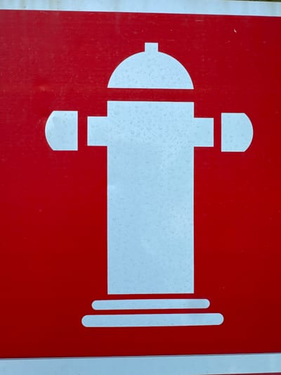 This is a close-up, overhead, eye-level, medium shot of a white fire hydrant symbol on a red background, likely a sign. The sign is wet, with many small water droplets covering the white symbol. The sign appears to be made of a material that has a slight texture. The bottom edge of the sign has a white border. No people, animals, or other objects are visible. The scene is likely outdoors, given the water droplets. The location in the city of Mejaniga, Italy, is not visually discernible from the image itself.