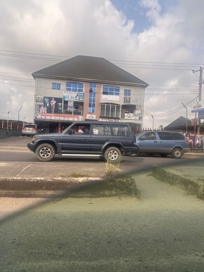 The image depicts a street scene in Elimgbu, Nigeria, under a partially cloudy sky. The central focus is a three-story commercial building with a dark grey hip roof and a light-colored facade featuring multiple blue-tinted windows and prominent vertical blue window sections. The ground floor houses several businesses, identified by signage including "ATLANTA STUDIOS," "DIAMOND SHOPPING MALL," "DG DE ABLE GOD," and "V.W. Fashion World."

On the paved road, a dark blue-grey Mitsubishi Pajero-style SUV is traveling from left to right. A male driver wearing a light-colored shirt and a front passenger are visible inside the vehicle. Behind this SUV, positioned further right, is a light blue-grey minivan. On the far left, partially obscured by a dark fence, a light-colored SUV is visible.

The foreground features a concrete curb separating the asphalt road from a raised, unpaved median containing sparse green vegetation. Overhead power lines are visible against the sky, extending across the upper portion of the frame. On the far right, a utility pole stands near a sign displaying "EYE LAND," adjacent to another building with a dark roof.
