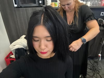 This image depicts an indoor scene within a hair salon in Herentals, Belgium. In the foreground, a young East Asian female, facing forward with her eyes closed and head tilted downwards, is seated. She has straight, dark hair parted in the middle, with a silver hair clip holding a section of her hair upwards. She wears a black, long-sleeved ribbed top and has visible pink lipstick and dark eye makeup. Positioned behind her, an adult female with light brown hair, wearing a black garment with ruffled sleeves and a silver smartwatch on her left wrist, is actively styling or treating the first subject's hair, holding a section with her right hand. The background features dark grey cabinetry, a black microwave oven positioned on a shelf, and an upper shelf containing multiple white mugs. To the left, a red plastic crate holds a light-colored padded jacket. On the right, a dark counter holds various items, and power cables along with a white power strip are visible on the floor.