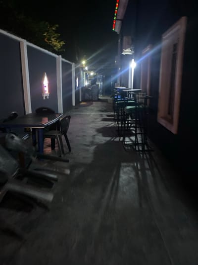 The image depicts an unoccupied outdoor commercial seating area at night in Igbessa, Nigeria. The scene is illuminated by multiple artificial light sources, producing lens flares and long shadows across the ground.
On the left, a grey perimeter wall with white vertical accents features at least one recessed, pointed arch-shaped alcove emitting a purple-pink light. Tree foliage is visible above this wall. In the immediate foreground, a dark square table is positioned with two dark chairs, while additional dark chairs are stacked or overturned on the concrete ground.
On the right, a dark-colored building facade displays at least two light-colored, rectangular window frames or decorative panels. Red and green lights are visible along the top right edge of this building. A row of high-top tables with associated high-back bar stools, featuring dark frames and green upholstered seats, extends into the mid-distance along this facade.
The ground surface is dark grey, appearing to be concrete, with some reflective areas possibly indicating dampness. No individuals are present in the frame.