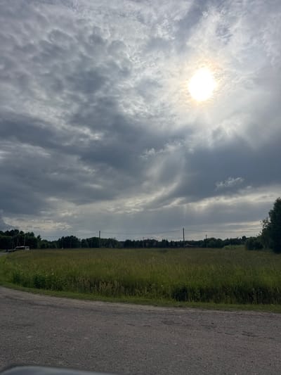 The image depicts an overcast sky with the sun peeking through, creating visible rays. Below the sky, a field of tall green grass stretches out, bordered by a line of trees and utility poles in the distance. A dirt road curves into the foreground from the left. The scene is set in a rural area, possibly on the outskirts of a town or village, given the presence of utility poles and scattered structures visible within the treeline. The overall impression is one of a natural landscape under a dynamic sky.