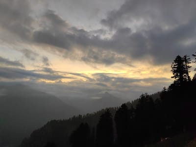 The image depicts a mountain landscape at dusk, likely in Siumsa, India. The sky is filled with dramatic, dark clouds illuminated by a band of golden light breaking through on the horizon. Silhouetted against this dramatic sky are the dark, jagged shapes of pine trees lining a steep mountainside in the foreground. In the middle ground, layers of mist or fog obscure distant, darker mountain peaks. The overall atmosphere is moody and serene, suggesting a natural, remote environment.