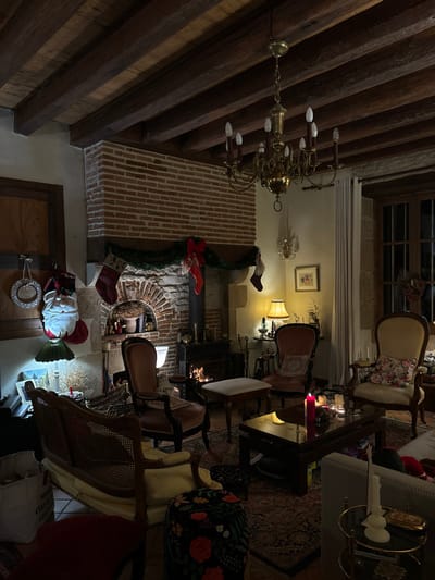 This image captures a warm and inviting, antique-style living room, likely in a traditional European home, decorated for Christmas. The setting is in Étréchy, France.

The most prominent features are the exposed dark wooden beams on the ceiling, a large brick fireplace, and a wood-burning stove with a visible fire, providing a cozy glow. The fireplace is adorned with green garlands, a red bow, and two Christmas stockings hanging from the mantelpiece. Another Christmas stocking or decoration is visible above the mantel, and a Santa Claus figure along with a wreath-like decoration are near a wooden door on the lower left.

The room is furnished with several classic upholstered chairs, some with cane backs, arranged around the fireplace and various side tables. A brass chandelier hangs from the ceiling, contributing to the ambient lighting. Other light sources include table lamps and wall sconces, creating a soft, warm illumination throughout the space. A patterned rug covers the floor. Through a window, hints of darkness outside suggest it is evening or night. A framed picture hangs on the wall. The overall atmosphere is one of comfort, tradition, and festive cheer. No discernable text is visible in the image.