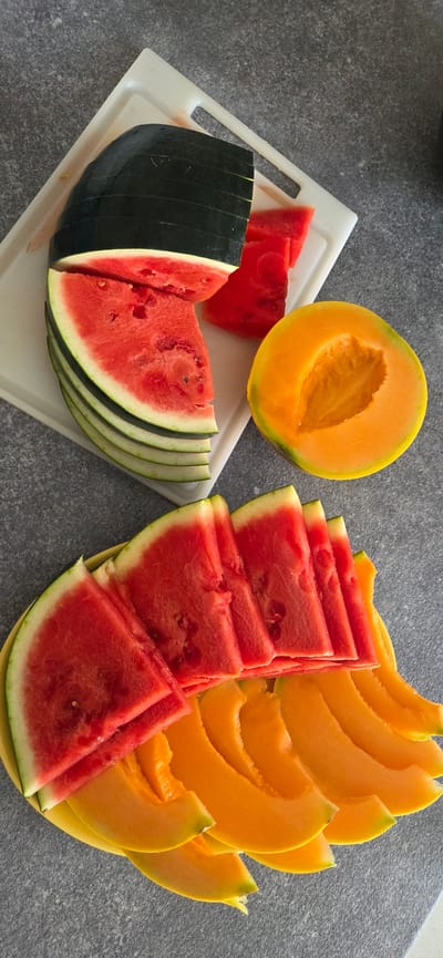 A high-angle, close-up shot shows a collection of sliced fruits, specifically watermelon and cantaloupe, arranged on a white cutting board and a yellow plate. The fruits are laid out on a speckled gray countertop. In the upper left, a partially sliced dark green watermelon is visible, with several fanned red slices and two smaller triangular pieces beside it on the cutting board. To the right of the cutting board, a halved cantaloupe with bright orange flesh and a scooped-out center rests on the countertop. Below, on the yellow plate, there are rows of fanned watermelon slices, overlapping with curved slices of cantaloupe. The scene is well-lit, highlighting the vibrant colors of the fruit. There are no people or animals present. The setting appears to be a kitchen or food preparation area. The location is not specified in the image.