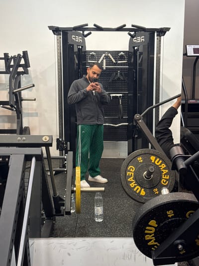 The scene depicts the interior of a gymnasium located in Plataniás, Greece. A male individual, positioned centrally, is capturing a mirror selfie using a smartphone held at chest height, partially obscuring his face. He is dressed in a dark grey zippered fleece jacket, green patterned athletic trousers with white vertical stripes, and white sneakers. Behind him, a large black multi-station cable machine or power rack, with the inverted text "GES" visible on its frame due to the reflection, stands against a white wall. This equipment features various handles and pull-up bars. To the left, additional black gym equipment frames are present. In the right foreground, part of a weight-loaded exercise machine, possibly a leg press, is visible, loaded with black and yellow circular weight plates labeled "GEARUP" and marked with "15" (likely kilograms). A clear plastic water bottle rests on the black speckled rubber gym floor near the man's feet. A partial reflection of another person's leg, clad in dark athletic wear, is visible on a reflective surface associated with the equipment in the right foreground.