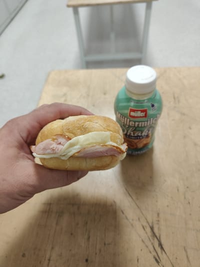 A hand holds a small sandwich with ham and cheese on a wooden surface. To the right of the sandwich is a bottle of Müller Milchshake. The background is out of focus and appears to be an indoor setting with a light-colored floor and a light-colored structure with a wooden shelf. The location is Łomna-Las, Poland.