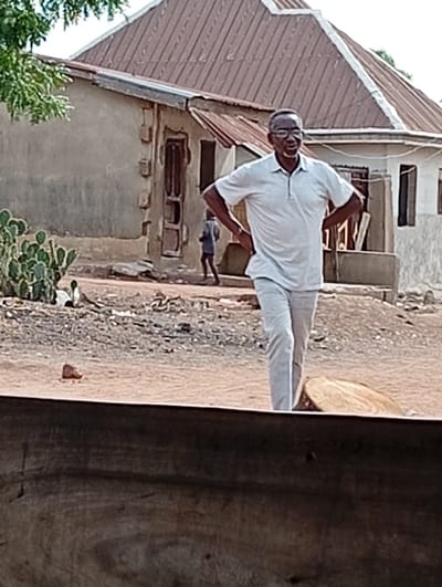 In Gboko, Nigeria, an outdoor scene unfolds on a dry, reddish-brown dirt path. A middle-aged to elderly man, wearing glasses, a light-colored short-sleeved polo shirt, and matching trousers, stands in the foreground with his hands on his hips, smiling. To his immediate right on the ground is a large, light-colored wooden log. Behind him, a small child in a dark shirt and shorts walks away from the viewer, moving towards the left. The background features two single-story buildings with reddish-brown corrugated metal roofs and light-colored, weathered walls. The building on the left has a wooden door with decorative trim. A green tree branch is visible in the upper left, and a cluster of cactus plants grows near the left background building. The ground is dusty with scattered small debris.
