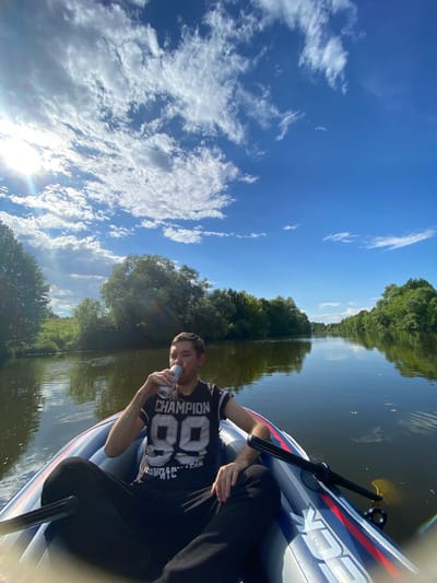 In the city of Chulki-Sokolovo, Russia, a man sits in an inflatable boat on a calm river, drinking from a can. He is wearing a black t-shirt with a graphic design and black pants. The boat is partially filled with water. Lush green trees line the banks of the river, reflecting in the water. The sky above is a clear blue, dotted with white, fluffy clouds, and the sun is shining brightly, casting a lens flare into the image. A paddle rests on the side of the boat.