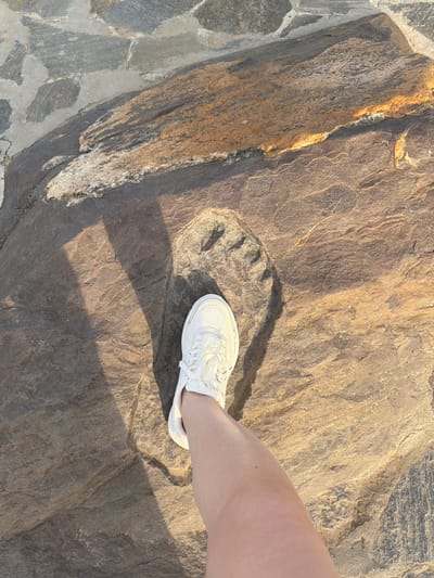 A person's leg and white sneaker are positioned over a natural rock formation with a distinct imprint. The imprint appears to be a footprint, with clear toe and heel definition. The rock itself is textured and varied in color, showing shades of brown, grey, and streaks of yellow and white, suggesting a geological formation. The background consists of similarly textured rocks, some of which are darker and appear to be part of a pathway or surrounding landscape. The lighting suggests it is daytime, with sunlight casting a shadow of the shoe and leg onto the rock. The scene appears to be outdoors, possibly in a natural park or historical site, given the presence of the imprint and the rugged terrain. There is no visible text in the image. The location context is Cherdyn, Russia.