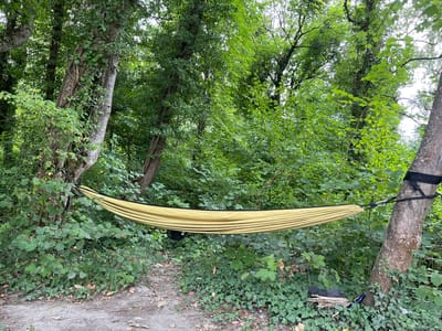 A yellow hammock is suspended between two trees in a wooded area. The trees are tall and covered in green leaves, and the ground is covered in dirt and fallen leaves. The hammock is empty and appears to be made of nylon. There is a black bag hanging from the hammock. The scene is peaceful and suggests a place for relaxation and nature enjoyment. The location is Étrembières, France.