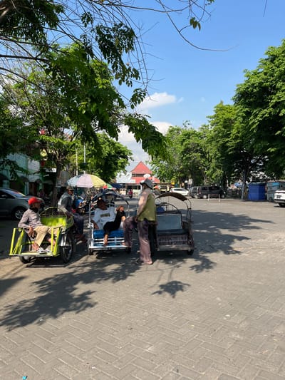 In Juwet Kenongo, Indonesia, on a bright, sunny day, several people are gathered near three-wheeled vehicles that appear to be rickshaws or modified motorcycles. The scene is set in an outdoor area with a paved ground surface, likely a street or market square, lined with trees providing dappled shade.
In the foreground, a man wearing a red cap and a plaid shirt sits in a bright yellow rickshaw. To his right, another man in a cap and a gray jacket is seated in a white and blue rickshaw, facing the viewer. A third rickshaw, darker in color, is positioned further back.
Several individuals are standing and interacting, seemingly engaged in conversation or waiting for passengers. One man, wearing a light green shirt and dark pants, stands near the second rickshaw. Another man, in a white shirt and dark pants, is seated in the same rickshaw, with his legs extended.
In the background, there are buildings with red roofs, and a variety of vehicles, including cars and what appears to be a black van. Various stalls or temporary structures, one covered with a blue tarp, are also visible, suggesting a marketplace or transport hub. The overall atmosphere is relaxed and casual, characteristic of daily life in a local Indonesian setting.