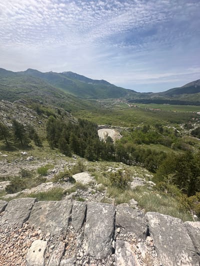 The scene depicts a mountainous landscape in Bukovica, Montenegro, as viewed from an elevated position. The sky shows patchy clouds. A winding road is visible in the distance, surrounded by dense forest and grassy terrain. The foreground consists of a stone retaining wall and rocky terrain, suggesting the vantage point is a constructed overlook. There are no visible people, subjects, or actions taking place. The primary subjects are the natural landscape, the road, and the constructed wall.