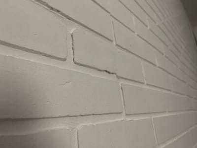 The image displays a close-up, angled view of a white painted brick wall. The focus is on a noticeable crack that runs horizontally across the mortar joint between two bricks, and then vertically down the face of one brick. The texture of the brick and mortar is rough and uneven, indicative of a painted surface. The lighting suggests an indoor environment, possibly a building in Izobilnyy, Russia, though no specific landmarks or identifying features of the city are visible in this cropped image. There are no people or other objects present.