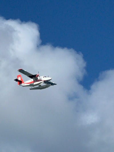 A twin-engine seaplane, predominantly white with red accents, is captured in mid-flight against a backdrop of blue sky and white clouds. The seaplane has floats beneath its fuselage, indicating its capability for water landings and takeoffs. The registration number "8Q-TRA" is visible on the tail fin. The overall scene suggests a clear day with scattered cloud cover, typical of tropical weather. The image is a photograph taken from below, looking up at the aircraft. The Malé city, Maldives, context suggests this type of seaplane is commonly used for inter-island transport and tourism in that region.